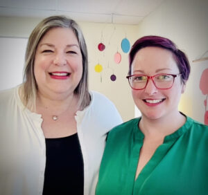 Picture of two female presenting people. One is wearing a black shirt with a white cardigan, the other is wearing a green shirt and has red glasses. 